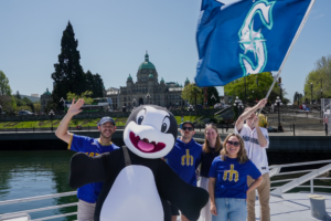 Seattle Mariners fans pose with Clippy the Orca on the Victoria Clipper in Victoria’s Inner Harbour, with the BC Parliament Buildings behind them.